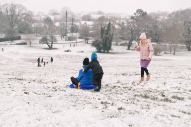 Küçük bir çocuk, annesini karda kızağın üstünde otururken itiyor. Anne, oğlu ve kızıyla dışarıda vakit geçiriyor.