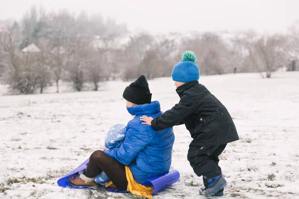 一个小男孩推着他的妈妈坐在雪地里的雪橇上 妈妈在外面陪着她的儿子和女儿 — 图库照片