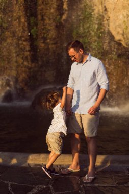 Genç baba, küçük oğluyla birlikte arka planda duruyor.