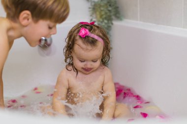 Küvette süt, portakal, sarı meyveler ve gül yapraklarıyla oturan tatlı küçük kız. Yıkanmak ve bir çocukla oynamak. Kıvırcık. Yan taraftaki çocuk..