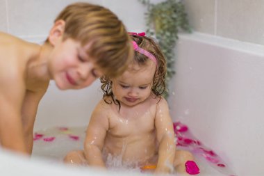 Küvette süt, portakal, sarı meyveler ve gül yapraklarıyla oturan tatlı küçük kız. Yıkanmak ve bir çocukla oynamak. Kıvırcık. Yan taraftaki çocuk..