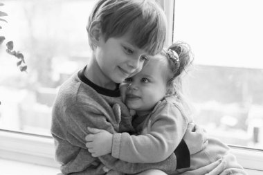 Mutlu çocuklar pencere eşiğinde oturup sarılıyorlar. Küçük kız ve oğlan. Siyah ve beyaz.