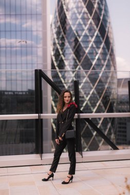 Klasik siyah takım elbiseli şık bir iş kadını. Güzellik. Büyürken. Arka plandaki cam binalar. Gökyüzü terası. Gökdelenler.