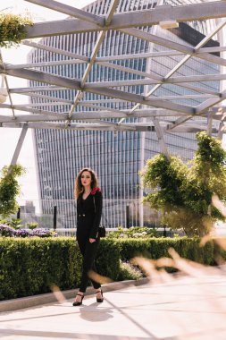 Klasik siyah takım elbiseli şık bir iş kadını parkta yürüyor. Güzellik. Büyürken. Arka plandaki cam binalar. Gökyüzü terası. Gökdelenler.