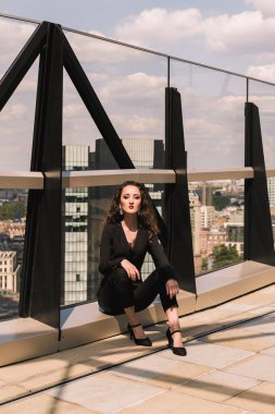 Klasik siyah takım elbiseli şık bir iş kadını. Otur bakalım. Güzellik. Arkaplan binaları, şehir manzarası. Gökyüzü terası. Gökdelenler.