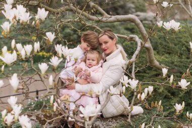 Genç anne, bahar parkında oturan kızını ve oğlunu kollarında tutuyor. Gülümsüyor. Çiçek açan manolyanın arkaplanı.