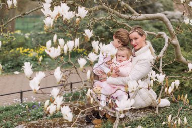 Genç anne, bahar parkında oturan kızını ve oğlunu kollarında tutuyor. Gülümsüyor. Çiçek açan manolyanın arkaplanı.