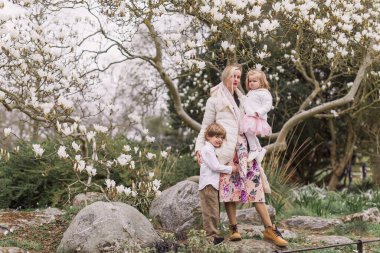 Genç bir anne bahar parkında kızını tutuyor. Ayakta. Gülümsüyor. Çiçek açan manolyanın arkaplanı.