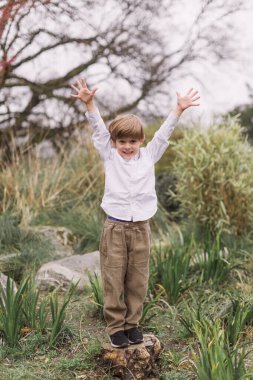 Beyaz gömlekli ve kadife pantolonlu küçük sarışın çocuk. Kollar havada duruyor. Açık havada hem de. Park et. Manolya çiçekleri.