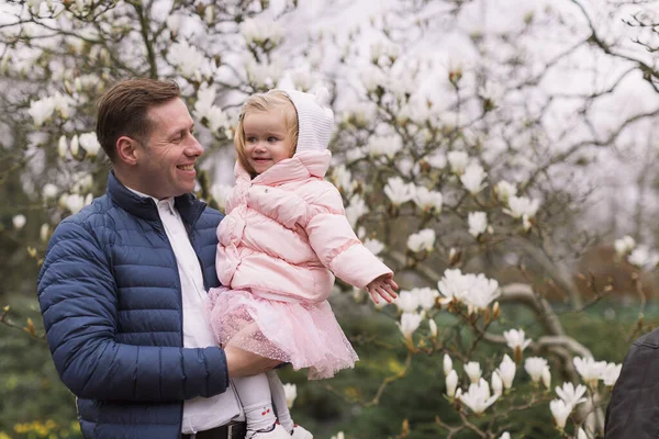 幸せな若い父親が娘を抱いている 暖かい服を着て 背景開花マグノリア — ストック写真