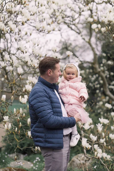 幸せな若い父親が娘を抱いている 暖かい服を着て 背景開花マグノリア — ストック写真