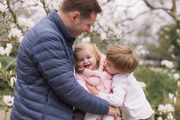 弟が抱きかかえると幸せな若い父親が娘を膝の上に抱えている 暖かい服を着て マグノリアの花の背景 — ストック写真