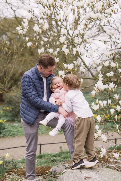 弟が抱きかかえると幸せな若い父親が娘を膝の上に抱えている 暖かい服を着て マグノリアの花の背景 — ストック写真