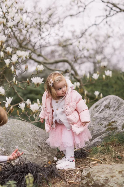 ピンクのスカートとジャケットのかわいいブロンドの幼児の女の子 屋外に立つ マグノリアの花の背景 — ストック写真