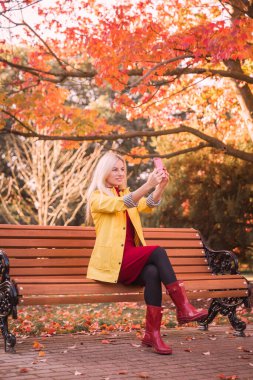 Genç sarışın kadın bankta otururken selfie çekiyor. Sonbahar parkında.