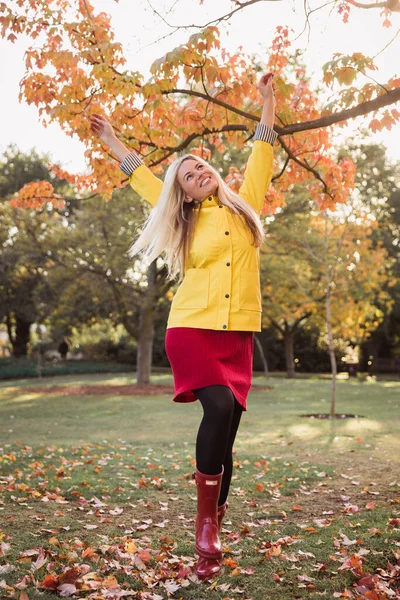 Young blonde woman in a yellow jacket having fun in the park. Standing. Autumn park background.