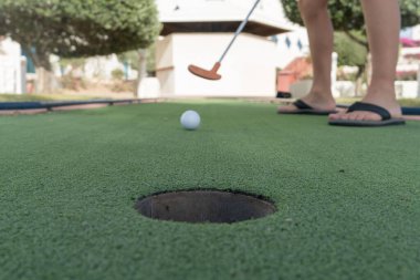 Küçük bir çocuğun ayakları minyatür bir golf deliğinde ya da küçük bir golf deliğinde top merkezde, dışarıda suni yeşil bir alanda kapanıyor..
