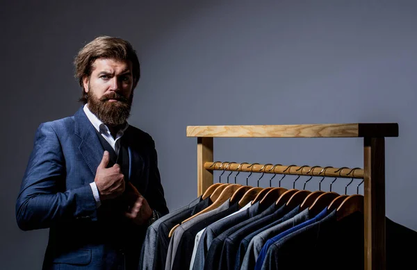 Male suits hanging in a row. Men clothing, boutiques. Tailor, tailoring. Stylish mens suit. Man suit, tailor in his workshop. Handsome bearded fashion man in classical costume suit