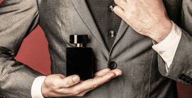 Elinde parfüm şişesi tutan bir adam. Takım elbisenin arka planında erkek parfümü var. Parfüm kokusu. Moda kolonya şişesi. Resmi takım elbiseli bir adam, bir şişe parfüm, yakın plan. Erkekler parfüm