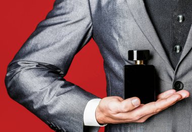 Elinde parfüm şişesi tutan bir adam. Takım elbisenin arka planında erkek parfümü var. Parfüm kokusu. Erkek parfümü. Moda kolonya şişesi. Resmi takım elbiseli bir adam, bir şişe parfüm, yakın plan.
