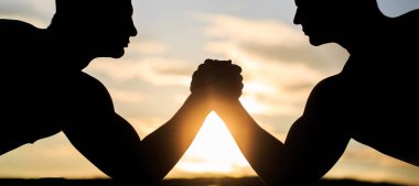 İki adam bilek güreşi yapıyor. Güçle yarışan ellerin silueti. Rekabet, meydan okuma, güç karşılaştırması. Gün batımı, gün doğumu. Rekabet, erkek bilek güreşine yakın çekim. Güç ve silah ölçen adamlar.