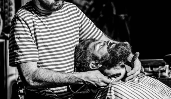 Bearded male sitting in an armchair in a barber shop while hairdresser shaves his beard with a dangerous razor. Barber shaving a bearded man in a barber shop.