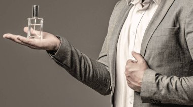 Resmi takım elbiseli bir adam, bir şişe parfüm, yakın plan. Parfüm kokusu. Erkek parfümü. Moda kolonya şişesi. Elinde parfüm şişesi tutan bir adam. Erkek parfümü, takım elbise arka planında