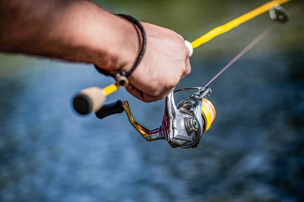 Pescador mano sosteniendo caña de pescar con carrete. Carrete de pesca. Caña de pescar con carrete de cuerpo de aluminio. Equipo de pesca. Suministros y equipo de pescado. Carrete de pesca primer plano en el fondo del río — Foto de Stock