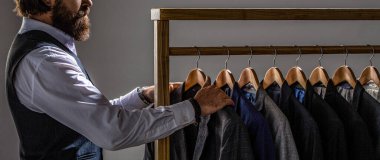 Klasik kostüm giymiş yakışıklı sakallı moda adamı. Erkek takımları peş peşe asılıyor. Erkek kıyafetleri, butikler. Erkek takım elbiseli, terzi de kendi elbisesinde. Terzi, terzi.