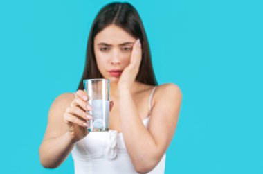 Kız hap alıyor, bir bardak su tutuyor, mavi üzerine izole edilmiş. Kadın baş ağrısına karşı ilaç alıyor. Esmer, bir bardak suyla hap alıyor. Baş ağrısını dindirmek için uyuşturucu alan bir kadın..