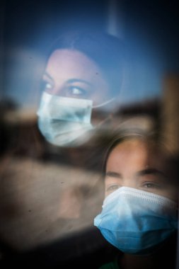 Bir anne ve oğlunun cerrahi koruyucu maskeler taktığı görüntüsü, pencerenin yanında, evlerinin içinde. Coronavirus COVID-19 salgını için kavramsal görüntü. 