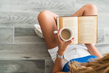 Entelektüel genç kız kitap okurken yerde oturup bir fincan kahve içiyor..