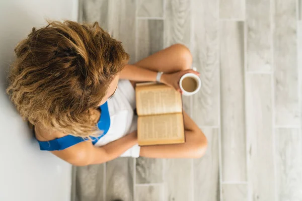 La joven intelectual se sienta en el suelo y bebe una taza de café, mientras lee un libro. —  Fotos de Stock