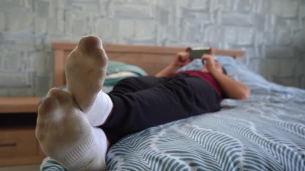 Мужчина в грязных белых носках лежит на кровати и играет по телефону — стоковое видео
