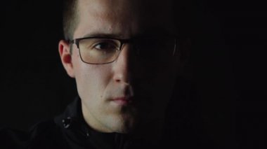 Gözlüklü adam loş ışıkta kameraya bakıyor.