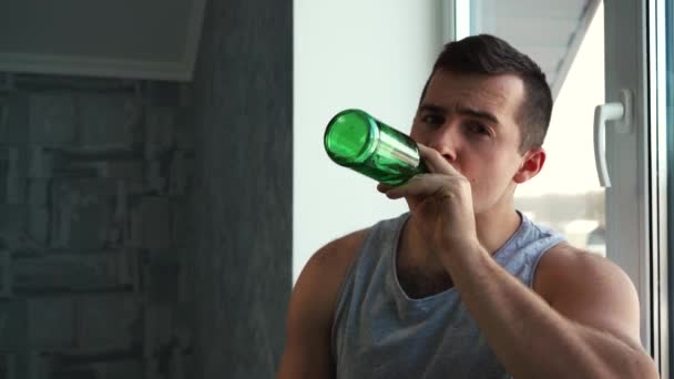 Человек пьет пиво из стеклянной зеленой бутылки, сидя у окна — стоковое видео
