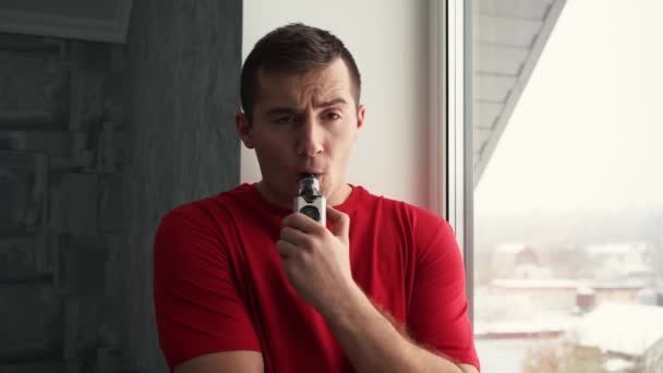 Un hombre con una camiseta roja fuma un vapor en casa parado junto a la ventana — Vídeos de Stock