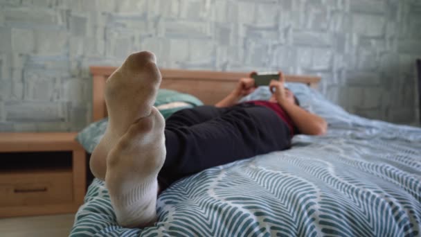 Een man in vuile sokken ligt op het bed en gebruikt de telefoon — Stockvideo