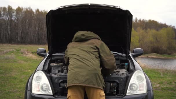 Un uomo che ripara un'auto distrutta in un campo vicino al fiume — Video Stock