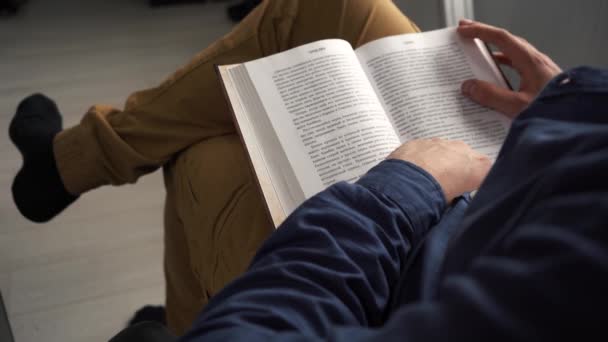 Egy férfi könyvet olvas, miközben egy széken ül a szobában. — Stock videók