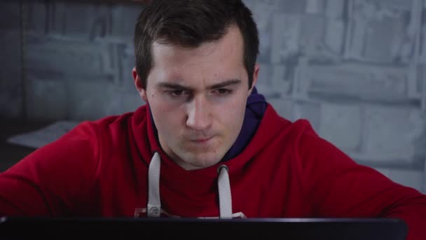 Ein Mann im roten Sweatshirt arbeitet, während er am Laptop sitzt — Stockvideo