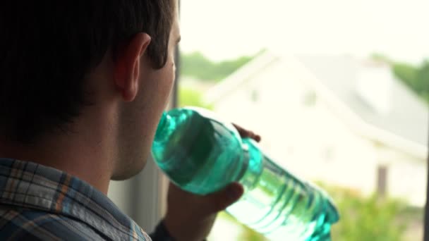 Un hombre bebe agua de una botella temprano en la mañana de pie junto a la ventana — Vídeo de stock