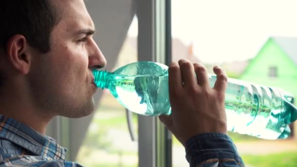 Un hombre bebe agua de una botella temprano en la mañana de pie junto a la ventana — Vídeo de stock