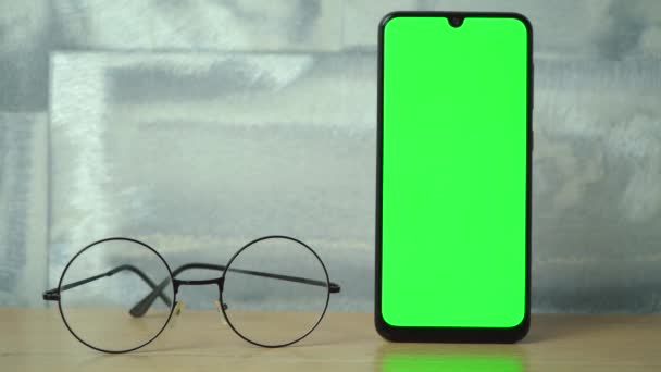 A phone with a green screen stands on a wooden table next to glasses – Stock-video