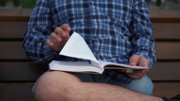 Egy férfi könyvet olvas, miközben egy padon ül. — Stock videók