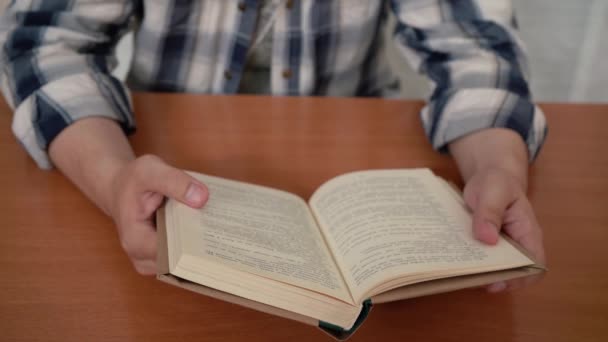 En man i skjorta läser en bok när han sitter vid bordet. — Stockvideo