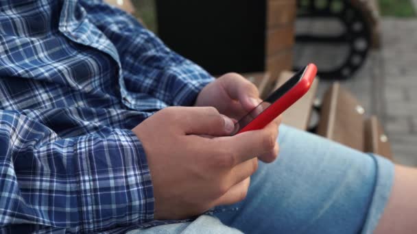 3.男人坐在公园的长椅上，手里拿着一部红色的手机 — 图库视频影像