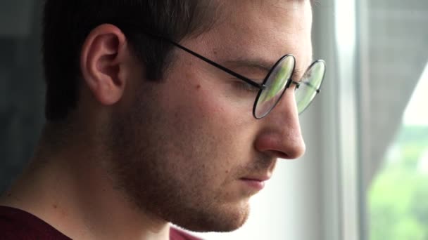 Человек в очках стоит у окна и смотрит — стоковое видео