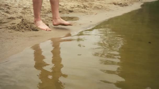 Een man loopt de rivier in met blote voeten op een zandstrand — Stockvideo