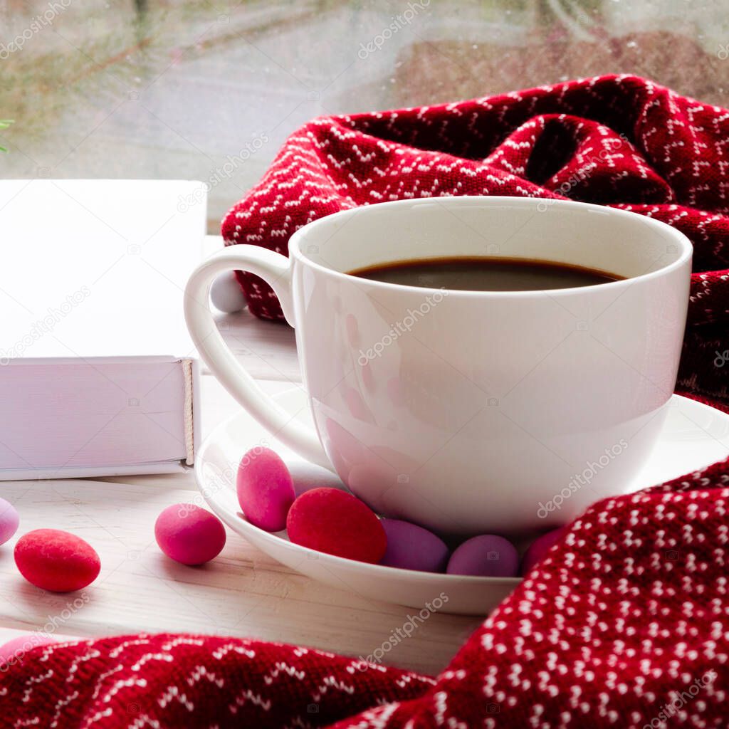 A cozy concept with a white cup of coffee, a red warm scarf, a white book and multi-colored candy on the windowsill